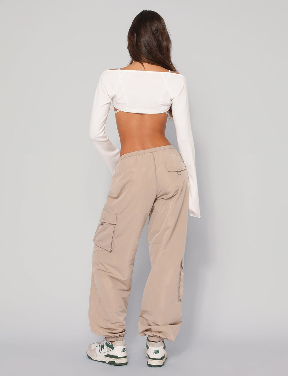 A person with long hair, facing away from the camera, wears a white open-back long-sleeve top, TIGERMIST's beige TRINITY PANT - NEUTRAL featuring an elasticated waistband, and white sneakers with green accents. The background is plain and light-colored.
