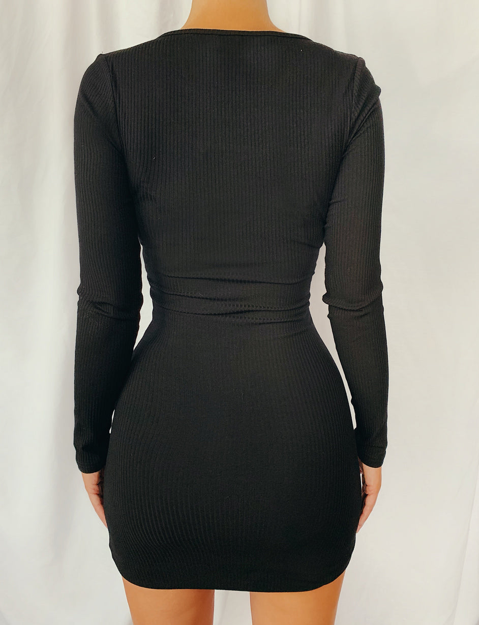 A person is wearing a form-fitting Aliyah Long Sleeve Dress - Black (Copy) from TIGERMIST, seen from the back. The dress has a ribbed fabric that accentuates the figure and reaches mid-thigh, featuring a scooped neckline. The background is a plain white curtain.