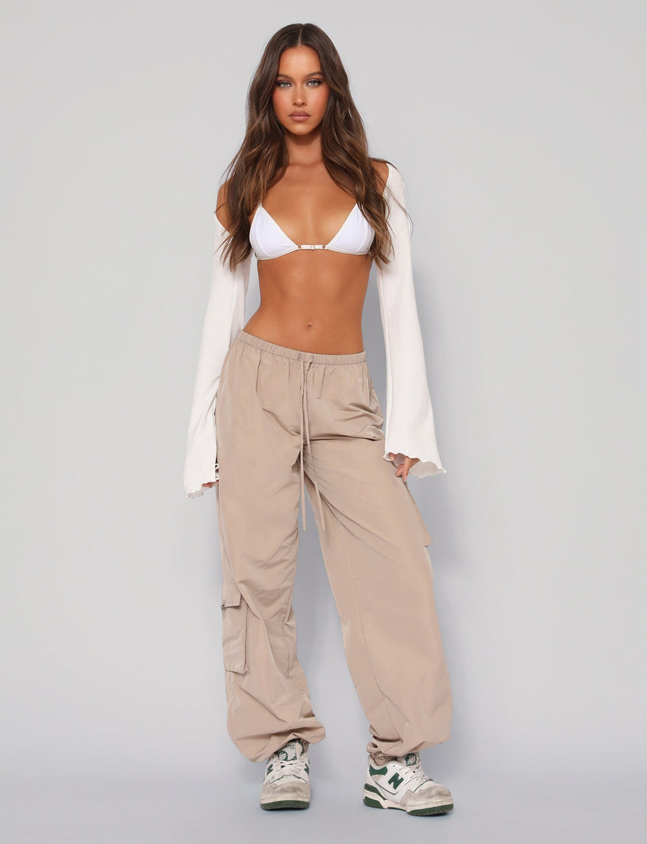 A woman stands against a plain backdrop wearing a white bikini top, a long-sleeve white shrug, and the TRINITY PANT in neutral beige from TIGERMIST. She has long, wavy brown hair cascading over her shoulders and feet adorned with white and green sneakers.