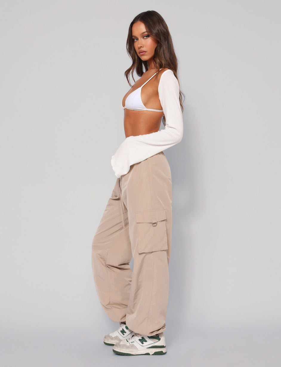 A woman stands confidently against a plain background, wearing a white bralette, long-sleeve white top, the soon-to-be-released TIGERMIST TRINITY Pants in beige with an elasticated waistband, and white sneakers with green details. She has long, wavy hair and adopts a casual yet stylish pose.