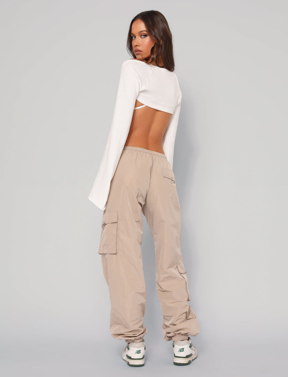 A woman with long, dark hair stands facing away from the camera. She is wearing a backless white long-sleeve top and the TRINITY PANT in neutral beige from TIGERMIST, featuring an elasticated waistband and multiple pockets. She is also wearing white sneakers. The background is plain and light-colored.